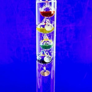 May include: A clear glass Galileo thermometer with four colored spheres inside. The spheres are red, yellow, green, and blue, each with a small metal tag indicating a temperature reading. The thermometer is set against a vibrant blue background.