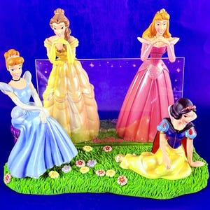 Puede incluir: Una figura decorativa con cuatro princesas de Disney: Cenicienta en azul, Bella en amarillo, Aurora en rosa y Blancanieves en amarillo. Las princesas posan sobre una base verde con flores, con un fondo púrpura.