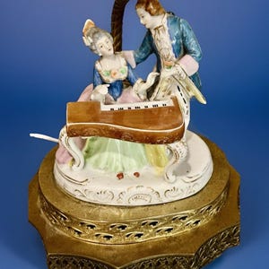 May include: A vintage porcelain figurine depicting a man and woman in period clothing playing a piano. The figurine features a brown piano, and the figures are dressed in blue, pink, and white. The base is gold-toned with ornate detailing.