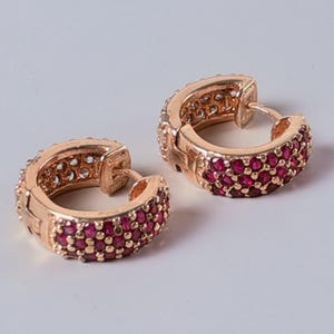 May include: A pair of rose gold hoop earrings featuring a hinged closure and a pavé setting of small, round, vibrant red stones covering the front half of each hoop. The interior of the hoops shows a polished rose gold metal finish.