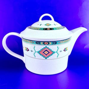 May include: White ceramic teapot with a lid, featuring a Southwestern geometric pattern in teal, orange, and black. The teapot has a curved handle and a spout. The lid has a small loop handle. The background is a solid blue.