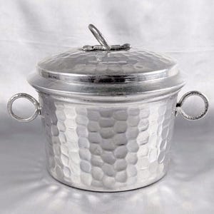 Puede incluir: Un recipiente de metal plateado martillado, con tapa y dos asas de bucle. La tapa tiene un pequeño asa. El recipiente tiene forma redonda y superficie texturizada. El diseño es sencillo y elegante.