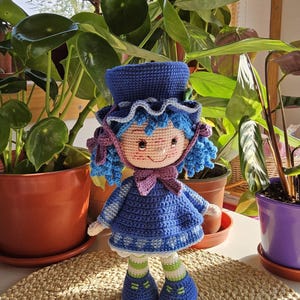 Puede incluir: Muñeca de ganchillo hecha a mano con cabello azul, un vestido azul y un sombrero a juego. La muñeca tiene una cara sonriente y está de pie sobre una estera tejida. La muñeca está rodeada de plantas en macetas.