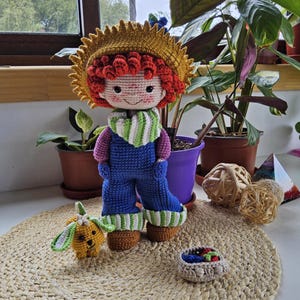 Puede incluir: Muñeca de ganchillo hecha a mano con pelo rizado rojo, un sombrero de paja amarillo y un peto azul. La muñeca tiene una bufanda a rayas verdes y blancas y botas marrones. También hay un pequeño perro de ganchillo y una cesta de bayas.