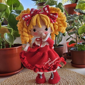 Puede incluir: Muñeca de ganchillo hecha a mano con pelo rizado amarillo y lazos rojos. La muñeca lleva un vestido rojo con ribete blanco y zapatos rojos a juego. La muñeca mide aproximadamente 25 cm de altura.