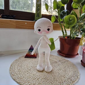 May include: A handmade, crocheted doll with a blank face, blush, and black eyes. The doll is white and stands on a round, woven mat. The doll's arms are outstretched, and it has large feet. The doll is a craft item.