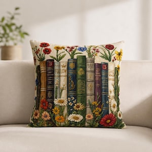 Può includere: Cuscino decorativo con un motivo di libri antichi e fiori. I dorsi dei libri sono di vari colori e il cuscino è decorato con margherite, papaveri e altri fiori. Lo sfondo è color crema.