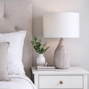May include: A bedroom scene featuring a white nightstand with a lamp, books, and a vase of greenery. The lamp has a textured, beige base and a white lampshade. A tufted headboard and bedding are also visible.