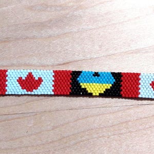 May include: A beaded bracelet featuring a design that combines the Ukrainian flag with the Canadian flag. The bracelet has a black background with blue and yellow Ukrainian flag elements, and red maple leaf symbols on a white background, with a silver clasp.