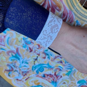 May include: A navy blue sleeve with gold flecks and a white lace cuff. The sleeve is resting on a guitar with a colorful floral design in yellow, blue, and red.