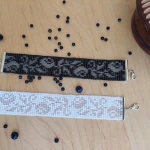 May include: Two woven bead bracelets featuring floral patterns. One bracelet is black with dark grey flowers, and the other is white with light grey flowers. Both have silver-toned metal end bars, lobster claw clasps, and short chain extenders.