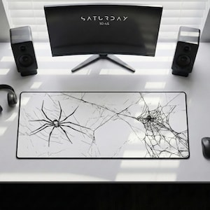 May include: A white rectangular mousepad featuring a black spider and web design. The mousepad has a black border and is placed on a white desk. A computer monitor displays the word "SATURDAY" and the time "10:45".