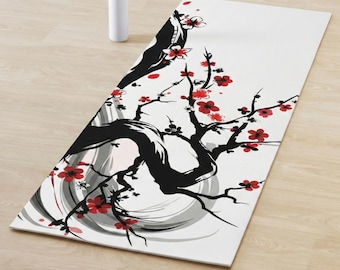 Op de afbeelding: Witte yogamat met een zwart en rood bloemmotief. Het kunstwerk toont een gestileerde boom met rode bloesems en een zwarte stam tegen een witte achtergrond. Een witte waterfles is ook zichtbaar.