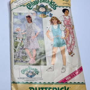Könnte beinhalten: Vintage Butterick Schnittmuster für Cabbage Patch Kids Outfits. Das Muster zeigt Illustrationen von Kinderbekleidungsdesigns, darunter Kleider und Overalls. Die Verpackung ist vergilbt und der Text "YES! IT'S EASY!" ist sichtbar.
