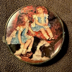 May include: A vintage round compact mirror with a painted image of two young girls in blue dresses, seated on a patterned surface with a black and white dog. The compact has a gold-colored metal frame.