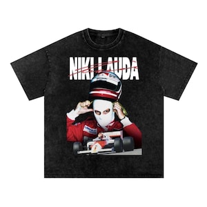 May include: Black t-shirt featuring a graphic of Niki Lauda wearing a racing helmet and balaclava, with a Formula 1 car in the background. The text "NIKI LAUDA" is printed across the top in white.