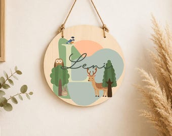Könnte beinhalten: Ein kreisförmiger Wandbehang aus Holz mit einem Waldmotiv, das einen Hirsch, eine Eule und einen Vogel zeigt. Der Name Leni ist in schwarzer Schreibschrift in der Mitte aufgedruckt. Das Design umfasst sanfte grüne und pfirsichfarbene Formen.