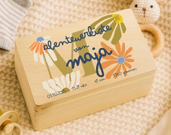 Könnte beinhalten: Eine hellfarbene Holzkiste mit floralen Designs und dem Namen "maja" in blauer Schrift. Die Kiste trägt den Text "Abenteuerkiste" und die Maße 47 cm und 3540 Gramm. Ein Schnuller und ein Stoffhase sind im Hintergrund.