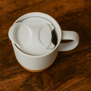 K&ouml;nnte beinhalten: Eine wei&szlig;e Keramik-Tasse mit hellgrauem Deckel und Henkel. Die Tasse hat einen Holzsockel. Der Deckel ist teilweise ge&ouml;ffnet und gibt den Blick ins Innere der Tasse frei. Die Tasse steht auf einer Holzoberfl&auml;che.