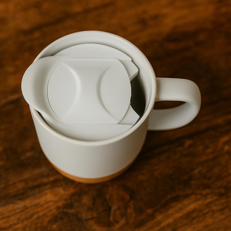 K&ouml;nnte beinhalten: Eine wei&szlig;e Keramik-Tasse mit hellgrauem Deckel und Henkel. Die Tasse hat einen Holzsockel. Der Deckel ist teilweise ge&ouml;ffnet und gibt den Blick ins Innere der Tasse frei. Die Tasse steht auf einer Holzoberfl&auml;che.