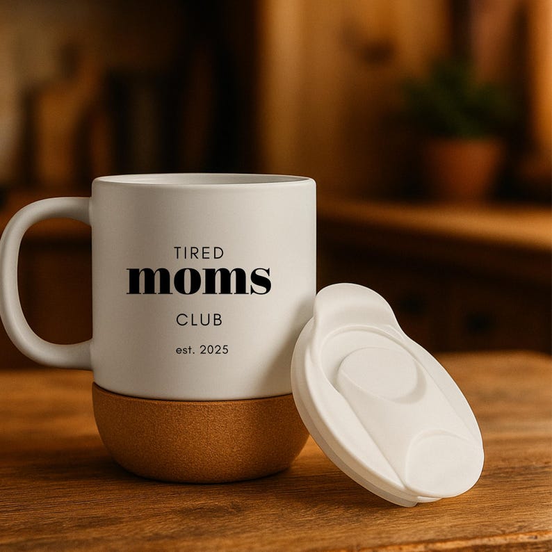 K&ouml;nnte beinhalten: Eine wei&szlig;e Keramik-Tasse mit dem Text "TIRED MOMS CLUB" und "est. 2025" in Schwarz. Der Boden der Tasse besteht aus Naturkork. Ein wei&szlig;er Deckel liegt neben der Tasse. Die Tasse steht auf einer Holzoberfl&auml;che.