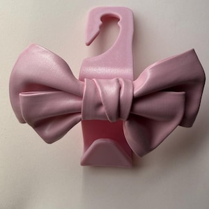 May include: A light pink bow-shaped hanger. The bow is made of a smooth material and is tied in the center. The hanger has a hook at the top for hanging clothes. The background is a plain white.