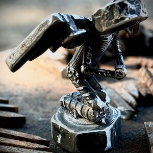 Puede incluir: Una escultura metálica artesanal detallada de un pájaro estilizado con las alas extendidas. La escultura es principalmente gris oscuro con acentos blancos, montada sobre una base hexagonal. La pieza tiene una estética industrial texturizada.