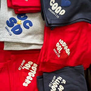 May include: Folded sweatshirts in red, grey, and navy blue, each with a unique design. The designs include text and heart shapes. The text includes names and the year 'C'28. The sweatshirts are neatly stacked.