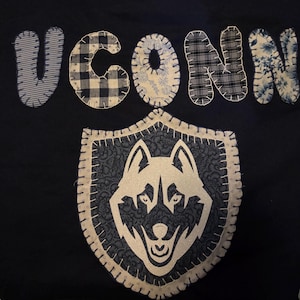 May include: Navy blue fabric with the word "UCONN" in patchwork letters. A shield-shaped emblem features a white husky dog design. The letters and emblem are stitched onto the fabric.
