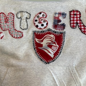 May include: A light gray hoodie with the word "KNIGHTERS" appliqued across the chest in various patterned fabrics. A red shield with a knight's helmet is also appliqued on the front.