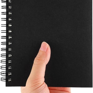 May include: A black spiral-bound notebook with a blank cover. The notebook is held by a hand, showcasing the clean, minimalist design. The wire binding is visible on the left side.