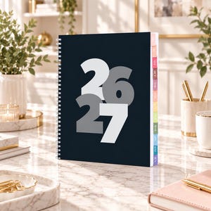 May include: A dark navy blue spiral-bound planner featuring the large white and grey numbers 26 and 27 on the front cover. The planner includes a side index with colourful monthly tabs labelled with three-letter abbreviations for each month of the year.