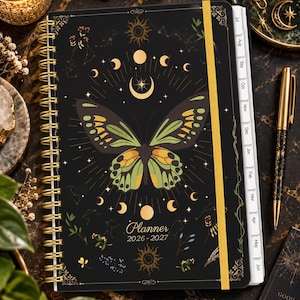 May include: A black spiral-bound planner for 2026-2027 featuring a central green and yellow butterfly illustration, gold sun and moon motifs, and celestial patterns. The cover includes a vertical gold elastic band and white monthly tab dividers.