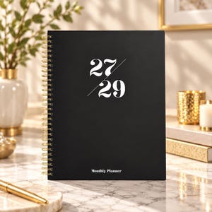 May include: A black spiral-bound monthly planner with gold-coloured wire binding. The front cover features the white text 27 over 29, separated by a diagonal line, and the words Monthly Planner printed in white at the bottom centre of the cover.