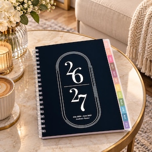 May include: A dark navy blue academic planner with white spiral binding. The cover features a white arched graphic with the numbers 26 and 27 stacked, and the text July 2026 - June 2027 Academic Planner. It includes colourful monthly tabs.