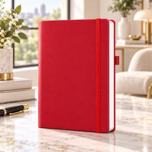 May include: A bright red hardback notebook with a vertical elastic closure band and a matching red fabric pen loop on the side. The notebook has a smooth, solid-coloured cover and white pages visible along the edge, standing upright on a surface.