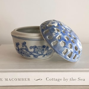 May include: A small, open ceramic container with a matching lid. The container is white with blue floral designs and a blue, perforated lid. The container is sitting on a white book titled "Cottage by the Sea".