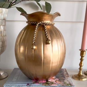 May include: A gold-colored decorative vase with a rope detail around the neck, sitting on a book titled "French Blooms." Two gold candle holders with pink candles are to the right. A pearlescent vase is to the left.