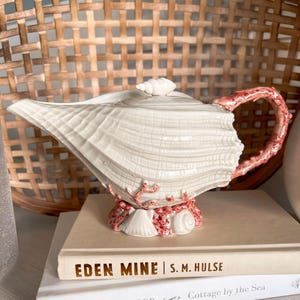 May include: A seashell-shaped teapot with a white glaze and coral-colored accents. The handle and base feature coral-like detailing. The teapot sits atop a stack of books, with the title "EDEN MINE" visible.