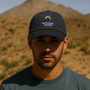 Puede incluir: Gorra de béisbol negra con el texto "EXPLORE MORE" y un gráfico de montaña amarillo y azul. La gorra la lleva una persona que viste una camiseta verde oscuro. El fondo es un paisaje desértico borroso.