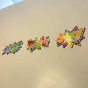 Puede incluir: Tres coloridos bocadillos de cómic con las palabras 'BISH', 'BASH' y 'BOSH!!' en un degradado arcoíris. Las burbujas tienen un diseño de estrella y están sobre un fondo blanco.