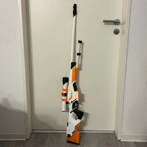 Könnte beinhalten: Ein Spielzeug-Scharfschützengewehr in Weiß, Orange und Schwarz lehnt an einer weißen Tür. Das Gewehr hat ein Zielfernrohr, einen langen Lauf und einen Griff. Das Farbschema ist hauptsächlich Weiß mit orangefarbenen und schwarzen Details. Das Design ahmt eine echte Schusswaffe nach.