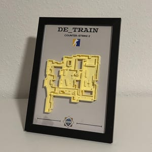 May include: A framed artwork featuring a yellow 3D model of a map, labeled "DE_TRAIN" and "Counter-Strike 2." The black frame surrounds a gray background with the map's layout. The artwork includes the Counter-Strike 2 logo.