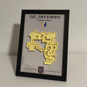 May include: Framed artwork featuring a yellow 3D map of a video game level, labeled "DE_INFERNO" and "COUNTER-STRIKE 2". The map is set against a light gray background, enclosed in a black frame.