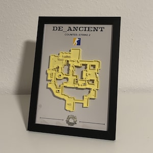 May include: Framed artwork featuring a yellow 3D map of a video game level, labeled "DE_ANCIENT" and "Counter-Strike 2." The map is set against a gray background within a black frame. A small logo is at the top.