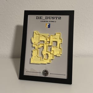 May include: Framed artwork featuring a yellow relief map of the "DE_DUST2" Counter-Strike 2 map. The black frame surrounds a gray background with the game's title and logo. A small emblem is at the bottom.