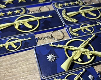 May include: A collection of rectangular keychains featuring blue backgrounds with dark blue camouflage patterns. The designs include yellow rifle silhouettes, gold-colored stars, and laurel wreath motifs. Each keychain is attached to a metal ring.
