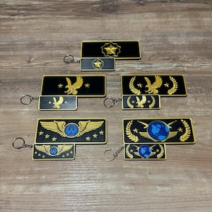 Könnte beinhalten: Schlüsselanhänger mit militärisch anmutenden Emblemen in Schwarz und Gold. Die Schlüsselanhänger zeigen verschiedene Designs, darunter Sterne, Adler, Flügel und einen Globus. Jeder Schlüsselanhänger hat ein rechteckiges Hauptteil und einen kleineren, passenden Anhänger.