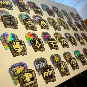 May include: A collection of stickers with various logos and text, including "NRG", "Red Canids", and "Lynn Vision Budapest". The stickers feature gold and black designs with ornate borders, some with holographic accents. The stickers are arranged on a white surface.