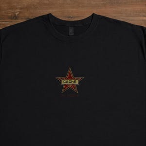 Könnte beinhalten: Schwarzes T-Shirt mit Rundhalsausschnitt und einem roten und goldenen Sterndesign. Der Stern zeigt das Wort "CACHE" und "CSGO" in einem rechteckigen Feld, darunter "do_cache". Das Shirt ist aus weichem Material.